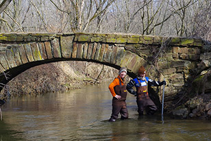 two female scientists stand in creek next to old bridge measuring